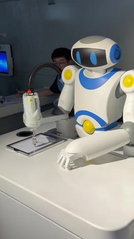 A robot arm with a pen attachment is drawing on a piece of paper placed on a white table. A person wearing glasses and a blue vest is visible in the background, seemingly operating or supervising the robot. Another person is partially visible on the left side of the screen. The environment appears to be an indoor setting, possibly a museum or exhibition. The robot is white with blue and yellow accents. The pen attachment is white and has a flexible grey tube connected to it.