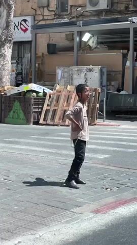 A young boy stands on a paved crosswalk, looking to his right with his hands on his hips and his mouth open in a smile. He is wearing a light brown t-shirt, dark pants, and dark shoes. He has a small black kippah on the back of his head. In the background, a man in a black hat and coat walks by from left to right, holding a phone to his ear. Further in the background, a man in a striped shirt pushes a shopping cart from right to left. The environment appears to be an urban street with buildings and some construction materials, including wooden pallets and plastic crates. The sun is shining, casting shadows on the ground.
