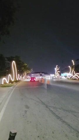 The video shows a road at night with decorative lights lining the side. Cars and motorcycles are driving on the road. There are also orange traffic cones in the road. The sky is dark and there are no visible stars or moon. The video is taken from the perspective of someone on a moving vehicle, likely a bicycle, looking forward.