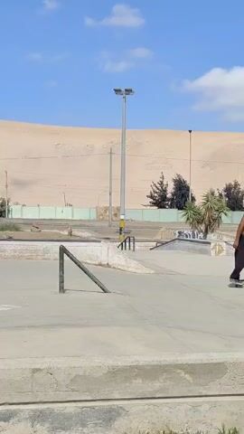The video shows a skateboarder in a skatepark. The skateboarder is wearing a black t-shirt, loose blue jeans, a white baseball cap, and white sneakers. He has dark hair and glasses. The skatepark is concrete with various ramps, rails, and other obstacles. There are streetlights and some vegetation in the background. Beyond the skatepark, there is a large sand dune under a bright blue sky with a few clouds. The skateboarder rides up to a long, low metal rail and successfully slides along it with his skateboard.
