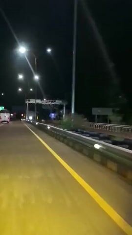 The video captures a nighttime view of a highway. Streetlights illuminate the road, casting a yellow glow on the asphalt. A yellow line marks the lane. In the distance, a toll booth structure is visible, with lights and what appear to be traffic signals. Several vehicles are present on the road, including cars and what looks like a van, all with their headlights on. The overall environment is dark, with the sky appearing black. There is a guardrail alongside the road.