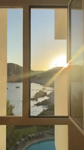 This media file captures a serene coastal scene at sunset in Andratx, Spain, viewed through a window. The primary subjects are the tranquil bay, dotted with several boats, and the lush, hilly coastline of the Mediterranean. The setting is a waterfront location, possibly a hotel or residential building, with a glimpse of a swimming pool and lounge chairs in the foreground.

The activity unfolding is a peaceful evening by the sea. The sun is setting, casting a warm, golden glow and creating sun flares that emanate from the horizon. The sky transitions from a clear, light blue at the top to a softer yellow and orange near the sun. The water is calm, reflecting the warm hues of the sunset. Small groups of people are visible on the beach and in the water, enjoying the fading daylight.

Notable details include the silhouette of the hills against the brightly lit sky, the rocky shoreline, and the evergreen trees that frame the bay. The overall emotion evoked is one of peace, relaxation, and natural beauty. The time of day is clearly sunset, indicated by the low angle of the sun and the warm lighting. The location cues of the Mediterranean coastline, the style of the buildings, and the vegetation strongly suggest a Spanish setting, specifically Andratx, as indicated by the provided context. There is no visible text in the media file.