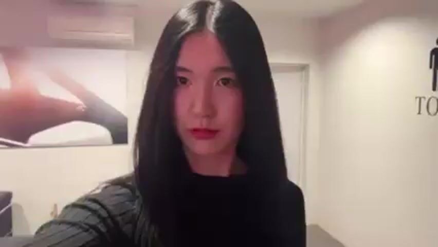 The video shows a woman with long, straight black hair and red lipstick. She is wearing a black sweater and is facing the camera. The background is a room that appears to be a waiting area or lounge. There is a sofa, tables, a beverage vending machine and a coffee machine. The room is well-lit. The woman is moving the camera slightly as she films herself.  There is a wall decoration showing a couple on the wall in the background.