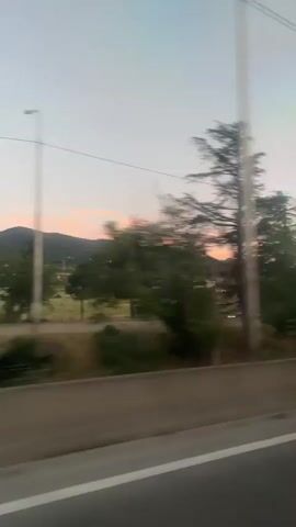 This is a first-person perspective video taken from inside a moving vehicle. The camera is pointed out the window to the right, capturing a landscape scene. The sky is a soft gradient of blue and pink, suggesting either sunrise or sunset. In the foreground, there is a concrete barrier with a white line, indicating the edge of a road or highway. Beyond the barrier, there are trees and vegetation. Some trees are blurred due to motion. In the background, there are rolling hills and mountains, with some structures visible, possibly houses or buildings. A street lamp is visible on the right side of the frame at one point. The overall impression is of a scenic drive through a rural or semi-rural area during twilight.