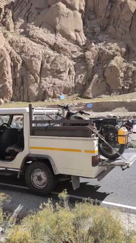 A white pickup truck with its doors open is parked on the side of a paved road. Two motorcycles are loaded in the bed of the truck, secured with straps. The truck is in a rocky, mountainous environment with sparse vegetation. A sign with text is visible on the right side of the road. The sky is clear and blue.