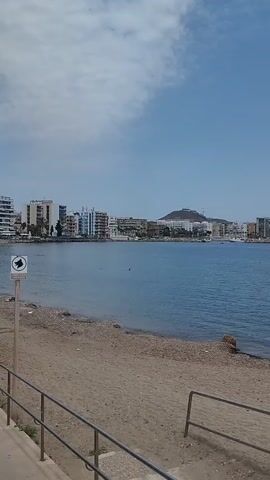 The video shows a view of a bay with a coastline. The water is a calm blue. On the left side of the frame, there are buildings lining the shore, including apartment buildings and what appears to be a hotel. There are also several boats anchored or sailing in the bay. In the distance, there is a rocky, tree-covered hill. The foreground shows a sandy beach with a metal railing. The sky is blue with a few wispy clouds. The video pans slowly across the scene from left to right.
