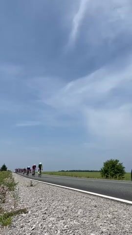 A cycling race is taking place on a sunny day with a bright blue sky dotted with wispy white clouds. The camera is positioned low to the ground, giving a dramatic perspective of the cyclists as they speed past. The road is asphalt, with a gravel shoulder on the left side. A line of cyclists, wearing colorful racing jerseys and helmets, are in motion. The lead cyclist, wearing a lime green jersey, is in sharp focus, while the others stretch out in a pack behind. The background consists of a flat, green field and a few scattered trees. The overall impression is one of speed, athleticism, and a competitive sporting event unfolding in an open, rural setting.