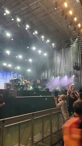 The video shows a concert stage with musicians performing. There are bright stage lights and smoke effects. In the foreground, a crowd of people watches the performance, some clapping and holding up their phones. The main focus is on a guitarist on stage who is moving and playing. The environment appears to be an outdoor or semi-outdoor venue with a large metal structure overhead supporting the stage and lighting.