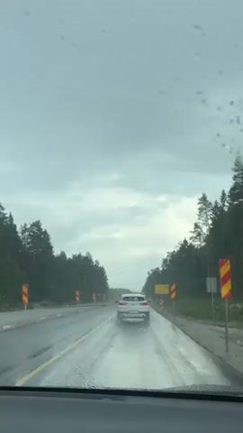 The video shows a view from inside a vehicle driving on a wet road under a cloudy sky. Raindrops are visible on the windshield. In front, a white SUV is driving ahead. The road is lined with trees on both sides, and there are construction barriers and warning signs along the right side of the road. The windshield wipers are visible in the foreground at 00:03.