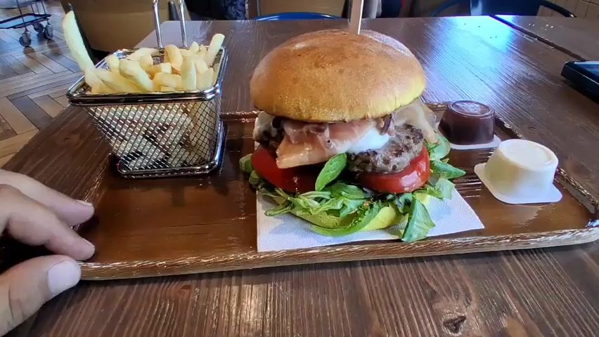 The video shows a close-up view of a hamburger and french fries on a wooden table. The hamburger is on a white paper liner, with lettuce, tomato, meat, cheese, and prosciutto visible. It is topped with a bun secured by a toothpick. To the left of the hamburger is a metal basket filled with french fries. To the right of the hamburger are two small white containers, one with a dark sauce and the other with a white sauce. A hand is visible in the lower left corner, reaching towards the french fries. In the background, blurry figures and furniture suggest a restaurant setting. The table surface is made of wooden planks.