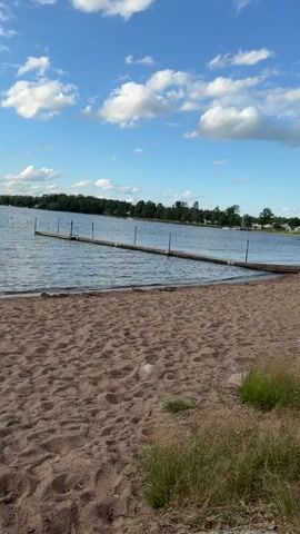 A sandy beach is in the foreground. A wooden dock extends into a body of water from the left side of the image. The sky is blue with white clouds. The water is rippling. Trees and houses are visible on the distant shore. The scene is lit by natural daylight.