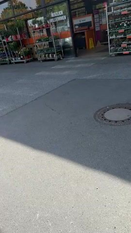 The video shows the entrance of a store. The sign above the entrance reads "EINGANG" which means "Entrance" in German. There are racks of flowers and plants outside the entrance. People are entering and exiting the store. One man is looking at the flowers. A woman is bending over, examining a plant. The store appears to be a garden center or hardware store with a garden section.