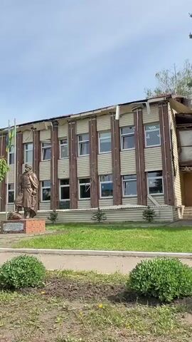 The video shows a two-story building with light-colored siding and brown vertical accents. The building has multiple windows on both floors. In front of the building is a bronze statue of a man on a pedestal, with the Ukrainian flag and another flag to its left. There is a small tree and some bushes in front of the building as well as a sidewalk and street. The building appears to have damage to the roof and siding. A billboard advertising food can be seen adjacent to the building. A tall evergreen tree stands to the side of the building. The sky is partly cloudy.