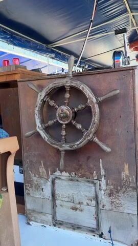 This media file features a close-up shot of a vintage wooden ship's wheel mounted on a weathered wooden panel. The wheel has a central brass hub and six spokes, each ending in a wooden handle. The wood of the wheel and the panel it's mounted on shows signs of age and wear, with a distressed, rustic appearance. Below the wheel, there's a section of the panel made of lighter-colored, rough-hewn planks.

The setting appears to be the interior of a boat, possibly a cabin or helm station, under a blue canvas awning. Red flashing lights are visible on a wooden structure to the left of the wheel, suggesting navigational or safety equipment. To the upper right, part of a boat's throttle control and what looks like a life preserver are visible. A portion of a wooden chair is on the far left.

The lighting suggests it is daytime, likely bright as indicated by the sunlight illuminating the scene and casting shadows. The overall impression is one of a classic, well-used vessel, possibly in a harbor or at sea. No people are visible in the frame. There is some faint, unreadable text on a white sticker-like object on the left side of the image.

The location context provided is Köyceğiz, Turkey, which is known for its lake and surrounding natural beauty, often explored by boat.