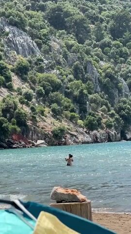 The video shows a scenic view of a cove or bay with clear turquoise water and a densely wooded, rocky hillside as a backdrop. Two people, seemingly a couple, are standing in the water, embracing each other. As the video continues, a young girl walks into the frame from the beach, wearing a colorful bikini and a pink headscarf. The camera is positioned on the beach, as evidenced by a partially visible beach blanket and what appears to be a weathered tree stump in the foreground. The day appears sunny and the overall atmosphere suggests a peaceful and relaxing beach outing.