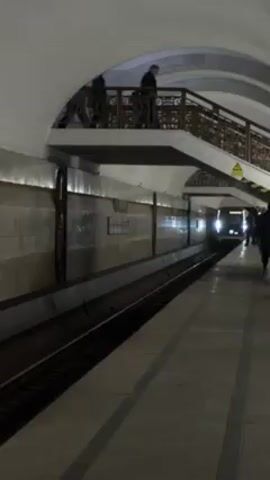 The video shows a subway train approaching a station. The station has a tiled floor, tiled walls, and an arched ceiling. There is a mezzanine level with a decorative railing, and people are visible on both levels. The train has its headlights on as it pulls into the station. People are waiting on the platform for the train.