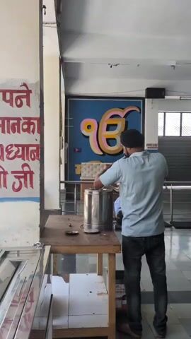 A man in a light blue shirt and black turban is pouring liquid from a metal pitcher into a larger metal container on a wooden table. A Sikh symbol is visible on a blue background behind him. Another person in an orange shirt is standing to the right, out of focus. Red text is visible on a white wall to the left.