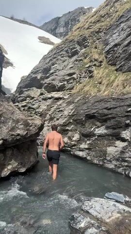 A shirtless man in dark shorts is wading through a shallow, rocky river in a mountain ravine. There is snow on the mountain above. Another man in a black shirt watches from the side.