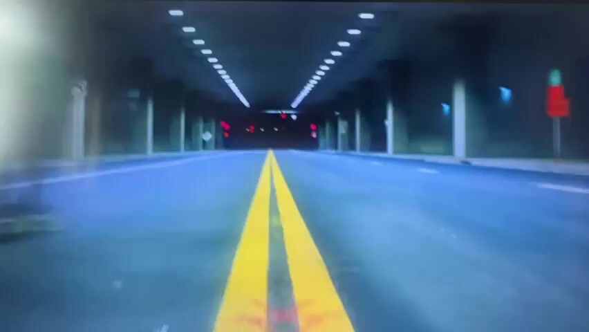 A low-angle shot follows a person skateboarding on a street, captured from the perspective of someone on a skateboard. The skater, wearing a green jacket and shorts, maneuvers their skateboard along a road marked with yellow lines. The environment appears to be an urban underpass or tunnel, characterized by concrete pillars and overhead lighting. The video then transitions to a blurred scene with pink and orange hues, featuring what seems to be people in a dimly lit room with wood-paneled walls and patterned wallpaper. This is followed by another blurry shot of two people skateboarding, this time with a blue-toned overlay, suggesting a different urban setting with brick structures in the background. The footage is characterized by its dynamic camera movement and use of color filters, focusing on the action of skateboarding and altered visual representations of indoor spaces.