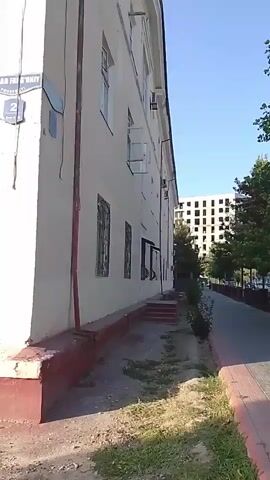 The video shows the exterior of a building. The building is light-colored with a red base. There are several windows visible, some with open shutters. A street sign with Cyrillic lettering and the number '2' is attached to the corner of the building. A sidewalk with a red and grey patterned border runs alongside the building. Trees and a partially constructed building are visible in the background. The sky is clear and blue. The video is shot from a low angle, looking up at the building.