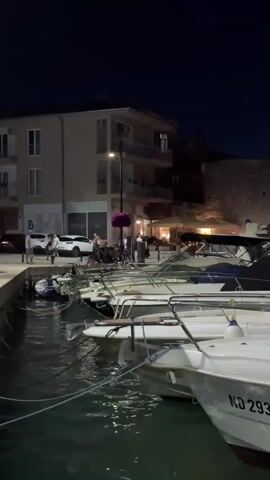 A nighttime scene shows a harbor with several boats docked in the foreground. Beyond the boats, a street runs alongside the water, lined with buildings. People are visible walking on the street and gathered near the boats. Lights from the buildings and streetlights illuminate the scene, reflecting on the water. One boat in the foreground has the license plate "ND 293".