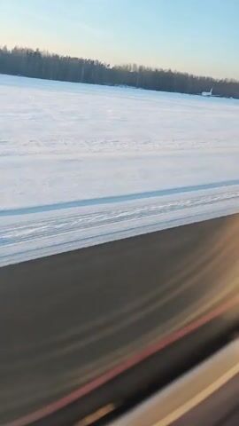 The video shows a landscape of snow-covered ground from the perspective of a plane window. In the distance, there is a forested area on the horizon, and a white airplane is parked on the snow. The sky is clear and blue. The camera is angled, showing a part of the plane's interior along the bottom edge of the frame. The video is somewhat shaky.