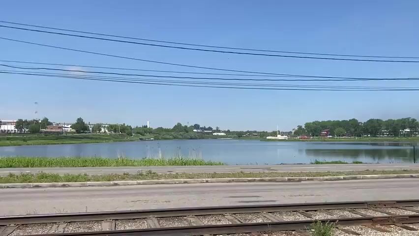 The video captures a serene daytime view of a large body of water, likely a lake or wide river, under a clear blue sky. In the foreground, railroad tracks run parallel to a paved road, which is separated from the water by a grassy strip. A tall utility pole with numerous power lines stands prominently on the right side of the frame, occasionally passing out of view as the camera moves.

Across the water, a distant shoreline is lined with various trees, beyond which several buildings and what appear to be industrial structures or tall towers are visible, suggesting a town or city in the background. The water's surface reflects the sky and distant elements, appearing calm with minimal ripples. Patches of reeds and other vegetation grow along the water's edge.

A green metal fence is positioned by the water, and near it, a solitary figure can be seen sitting on the bank. The overall impression is one of tranquility on a bright, sunny day. No text is visible in the video. The scene depicts a landscape that could be part of Usolye-Sibirskoye, Russia.