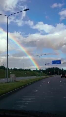 The video shows a view from a vehicle moving on a highway. A rainbow is visible in the sky to the left of the frame. The highway has multiple lanes, indicated by white arrows painted on the asphalt. Streetlights are positioned along the side of the road. A blue overhead sign with directional arrows is visible. Green grass and trees line the roadside. A dark-colored car is moving ahead in the right lane. Round red and white traffic signs are also visible. The sky is blue with white clouds.