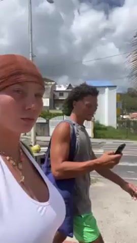 A woman wearing a white top and brown head wrap walks towards the camera, while a man in a tank top and green shorts walks behind her looking at his phone, then points. They are walking on a sidewalk next to a road. There are houses and trees in the background. The camera pans to show a view of the ocean and a palm tree. The sky is partly cloudy.