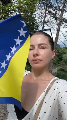 This is a selfie video of a young woman holding a flag of Bosnia and Herzegovina. She is outdoors with trees and foliage in the background, suggesting a natural setting. The woman is wearing a white top with black polka dots and a black bra underneath. She has her hair pulled back and is wearing sunglasses on top of her head. She raises the flag with her right hand and makes a kissing face towards the camera. The flag is clearly visible, featuring a blue field with a yellow triangle and a row of white stars. The lighting suggests it is daytime, likely late morning or afternoon, with bright sunlight casting shadows. The overall mood appears cheerful and patriotic, with the woman expressing pride in her country's flag. There is no visible text in the video. The location cues, combined with the flag, strongly indicate she is in Bosnia and Herzegovina, and specifically Jajce, given the provided context.