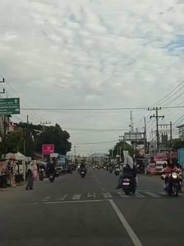 The video shows a street scene. There is a two-lane road with a pedestrian crossing in the foreground. Many motorcycles are driving on the road. On the sides of the road, there are buildings, trees, and power lines. Some people are walking or riding on motorcycles. The sky is cloudy.