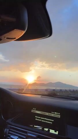 The video is shot from inside a car looking out through the windshield at a sunset. The sun is low on the horizon, casting a warm, orange glow across the sky and illuminating the clouds. There are mountains in the distance. The car's dashboard is visible in the foreground, with a display screen showing text in Russian. The text appears to be related to audio settings or Bluetooth connectivity. The overall impression is a peaceful and scenic moment of driving at sunset.