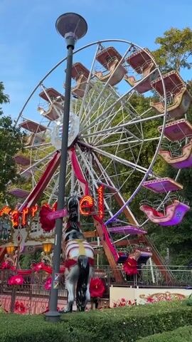 A carousel Ferris wheel is shown with colorful gondolas. A street lamp stands in the foreground, obscuring part of the wheel. A carousel horse is visible to the left. The Ferris wheel has red and gold accents.