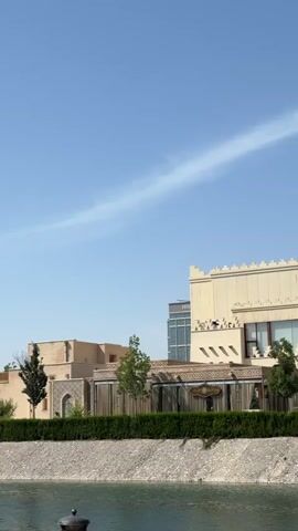 The video presents a daytime panoramic view of traditional Uzbek architecture situated along a tranquil body of water. The camera pans from left to right, revealing a complex of ornate buildings under a clear blue sky.

At the beginning, a large, light-colored building with crenellated rooftops and a modern structure (partially obscured) with the text "HILTON" visible in the background are shown. As the camera moves, more details of the traditional buildings come into focus, characterized by intricate tile work in blue, green, and white patterns, arched doorways, and decorative facades.

The buildings are separated from the water by a low, well-maintained green hedge and a stone-lined bank. Several mature green trees dot the landscape, providing shade and contrast to the light-colored structures. Towards the end of the pan, a decorative bridge with white railings connects different sections of the architectural complex, and a minaret-like tower with blue and white tile work becomes visible. The water reflects the clear sky, enhancing the serene atmosphere of the scene. The overall impression is one of cultural richness and architectural beauty in a peaceful setting.