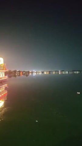 A large, multi-decked boat is illuminated with red and yellow lights at night. The boat is on a body of water, and its lights are reflected on the water's surface. The sky is dark, and there are distant lights on the shore.