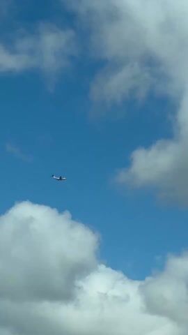 A military transport aircraft, identified as an Airbus A400M Atlas, is captured flying across a bright blue sky in Broadland, United Kingdom. Initially, the grey aircraft is clearly visible against a backdrop of scattered white cumulus clouds, flying from left to right across the frame. Its distinctive high-wing design and T-tail are prominent. As the video progresses, the aircraft approaches a larger, more diffuse cloud bank. It then flies directly into this cloud, gradually becoming obscured. By the end of the clip, only a dark, hazy silhouette of the plane is visible through the dense cloud cover. The sound of the aircraft's engines is audible throughout the daytime video, intensifying as it passes and then fading slightly as it enters the cloud.
