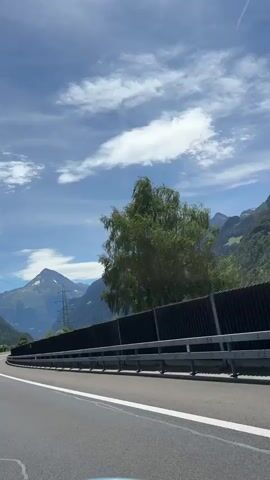 A vertical video shows a view from the passenger seat of a car driving on a highway. The highway is paved with asphalt and has white lane markings. On the left side of the highway, there are mountains, a truck, and a power line pylon. On the right side of the highway, there is a black sound barrier and trees. The sky is blue with white clouds. The video captures the car moving forward at a moderate speed.