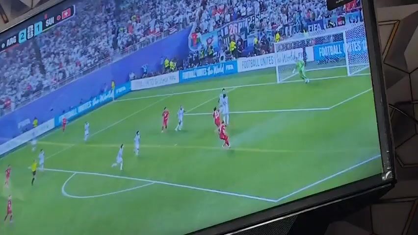 The video shows a television screen displaying a soccer match. The score on the screen reads ARG 2:0 SUI. Players in white and light blue uniforms are on one side of the field, while players in red uniforms are on the other. The goalkeeper in the red uniform is diving to save a shot. The crowd in the background is wearing white clothing. The television is black and has a mesh speaker grill at the bottom. The background behind the television is dark and features a geometric pattern.