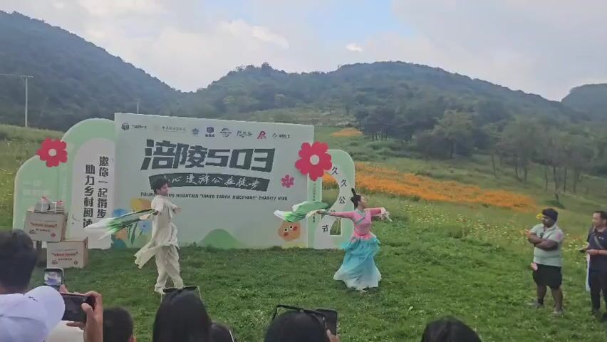 A group of people are gathered outdoors in a grassy field with hills in the background, possibly at an event. There is a large white banner with text and graphics, including the number 503. Two performers are dancing in front of the banner; one is dressed in a white outfit and the other in a pink and turquoise outfit, holding a green and white fan. A few spectators are visible in the foreground, with some holding up phones to record the event. To the left of the banner, there are boxes with drinks. The overall atmosphere suggests an outdoor festival or charity event.