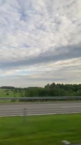 This video captures a scenic view from a vehicle traveling along a highway in the Knyazhytsky Selsoviet, Belarus. The scene is dominated by a vast expanse of lush green trees that form a dense forest lining the road. A highway with white lane markers stretches across the frame, indicating a journey through the countryside. Above, a sky filled with dramatic, billowy clouds suggests it might be late afternoon or early evening. The natural light filtering through the clouds creates a soft, diffused illumination, hinting at a cool or temperate climate. There are no people visible in the frame. The only man-made objects are the highway itself, a metal guardrail, and a solitary electrical pylon that stands tall against the horizon. The overall mood is tranquil and serene, evoking a sense of peaceful travel through a verdant landscape.