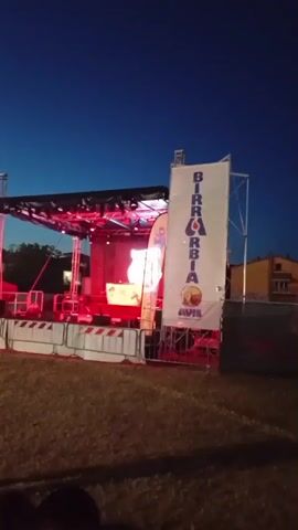 A temporary stage is set up on a grassy field at dusk. The stage is lit with red lights, and a large banner with the word "BIRRABIA" in large letters hangs on the right side. There are people sitting in the foreground, and the sky is dark blue with some faint clouds. The scene appears to be an outdoor event or festival.