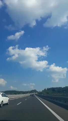 This is a vertical, eye-level video clip filmed from the perspective of someone in a car driving on a highway. The sky is a vibrant blue with scattered, fluffy white clouds, suggesting a clear and sunny day. The highway stretches out ahead, with multiple lanes marked by white lines.

The primary subject is the highway itself, with a few cars visible. In the foreground, a white car is in the left lane. Further ahead, other cars are visible in the distance, moving along the same highway. The setting appears to be a rural or semi-rural area, with a dense line of green trees bordering the right side of the highway, indicating a forest or wooded landscape. A metal guardrail separates the road from the trees.

The activity is driving on a highway. The time of day is likely daytime due to the bright sunlight and clear sky. The overall mood is one of travel and openness, with the expansive sky and the open road. No specific emotions are conveyed beyond the sense of movement and journey. There is no visible text within the frame. The location context of Skomielna Biała, Poland, suggests this highway is likely a part of the Polish road network.