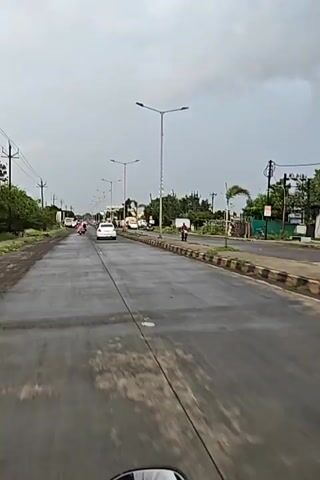 The video shows a long, straight, two-lane road with a concrete median in the center. The road appears to be wet, suggesting recent rain. There are streetlights lining the right side of the road. On the left side of the road, there are trees and vegetation. Several vehicles are visible on the road, including cars, motorcycles, and what appears to be a bus. People are seen riding on the motorcycles. The sky is overcast with dark clouds. The scene is captured from the perspective of someone riding a motorcycle, as indicated by the handlebars visible at the bottom of the frame. There are no obvious signs of distress or unusual activity.