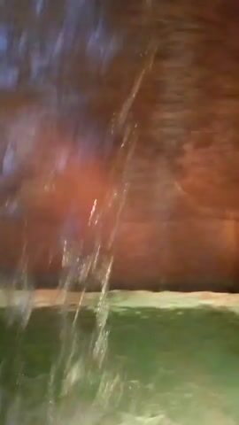 The video depicts the interior of a cave with water at the bottom. The walls are a dark, reddish-brown color. At the beginning of the video, water is seen trickling down the wall. The camera pans up slightly and then rests on a pool of green-tinted water at the base of the wall, before the video ends. The sounds of water splashing can be heard throughout the video.