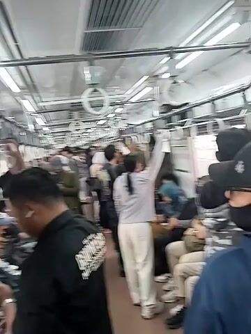 The video shows the interior of a crowded train car. The camera is positioned at eye level, facing down the length of the car. Numerous passengers are standing and sitting, with many holding onto overhead rails and straps for support. The lighting appears to be from overhead fluorescent fixtures. Several individuals are looking at their phones. The overall impression is one of a densely populated public transportation setting during what appears to be peak travel time. The color palette is muted, with predominantly neutral tones in clothing and the train's interior. The camera movement is minimal, suggesting a static recording.