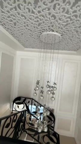 A drone camera pans down a grand staircase. The staircase has black wrought iron railings with a decorative scroll design. The walls are white and paneled. A large, modern chandelier with multiple silver orbs hangs from the ceiling. The ceiling itself has a white ornamental pattern. The camera moves past the chandelier and continues down the stairs, showing more of the ornate railings and paneled walls. The final shot shows a hallway with white doors and a white pillar.