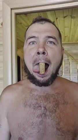 A shirtless man with a dark beard and hair stands facing the camera with a cork held in his mouth by his tongue. He is in a room with light wooden walls and a visible doorway behind him.