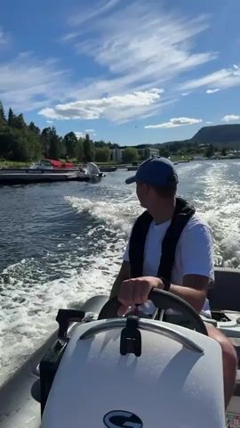 A man wearing a blue baseball cap and a white t-shirt with a black life vest is at the helm of a small inflatable boat, steering it through a body of water. The boat is creating a significant wake. In the background, along the shoreline, there is a dock with several boats tied up, some covered with red tarps. Further inland, on a grassy hill, a modern, multi-story house with large windows and balconies is visible. There are also several tall pine trees on the property. The sky is blue with scattered white clouds. The video appears to be a first-person perspective from the boat.