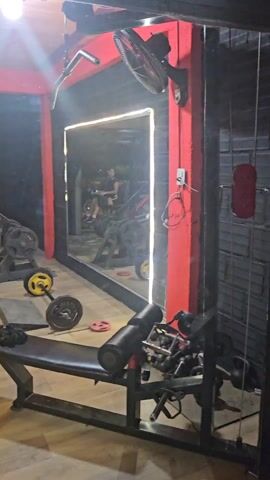 A person in a black workout outfit takes a selfie in a gym. They are wearing glasses, a black jacket, a black bra, black shorts, and gray sneakers. They hold a pink phone up to the mirror to take the selfie. There are workout benches and weights in the background. The gym is lit with overhead lighting. There is a mirror on the far wall. The wall trim is red. The gym floor is light wood grain.