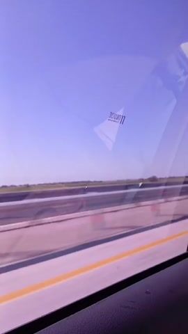 A blurred view from inside a vehicle shows a landscape of a road with some foliage in the distance, under a bright blue sky with purple hues. A sticker with the word "MIRACLE" is visible on the car window. The perspective suggests the vehicle is in motion, possibly on a highway. The overall impression is that of a travel snapshot.